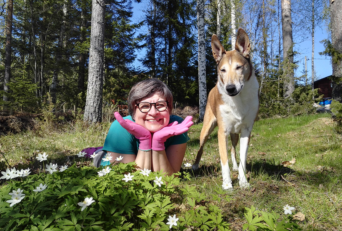 Anne Korhonen ja hänen koiransa valkovuokkojen ympäröimänä kesämökillä.