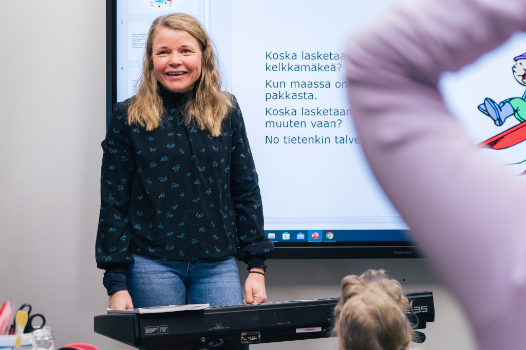 Vuokko Palokangas luokan edessä kosketinsoittimen kanssa säestämässä laulua, taustalla laulun sanat.