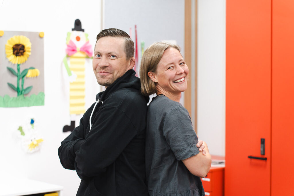 Jaakko Juvonen ja Anu Alenius-Taipalus nojaavat toistensa selkiin ja hymyilevät luokkahuoneessa.